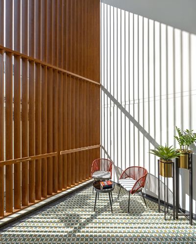 Sunlit balcony with wooden louvers, patterned tile floor, red woven chairs and brass planters