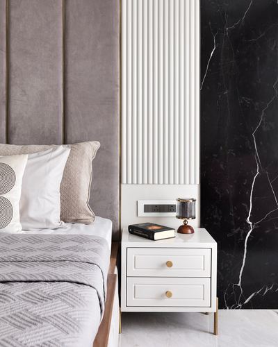 White bedside table with lamp and book beside grey velvet headboard and black marble wall panel