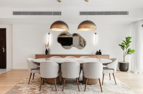 Dining room with oval marble table, grey upholstered chairs, and two brass dome pendant lights over a wooden sideboard