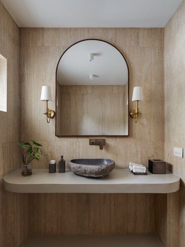 Arched mirror above a carved stone vessel sink on a floating beige counter, brass sconces flanking