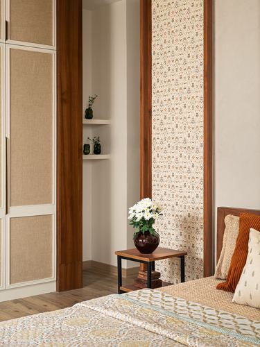 Bedroom corner with wood-framed patterned wallpaper panel, cane wardrobe, side table with white flowers, and bed with rust cushions