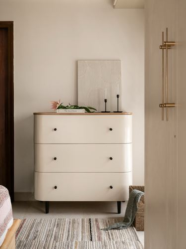 Cream rounded-corner chest of drawers with wooden top, candlesticks and textured art against beige wall