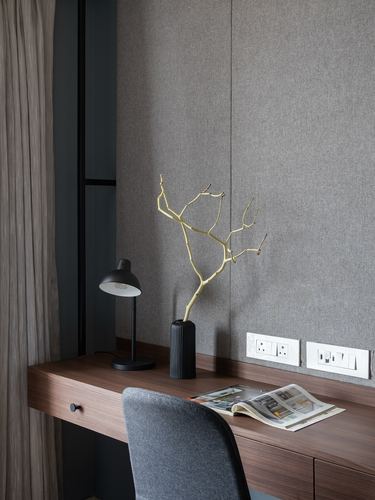 Wooden desk with black lamp, ribbed vase, dried branch, and grey upholstered chair against fabric-panelled wall