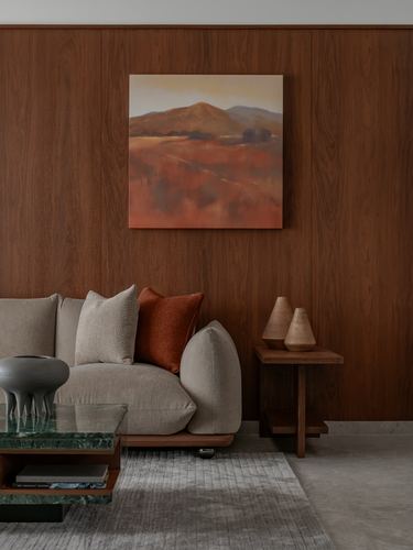 Beige sofa with rust cushion against wood-panelled wall with landscape painting and side table