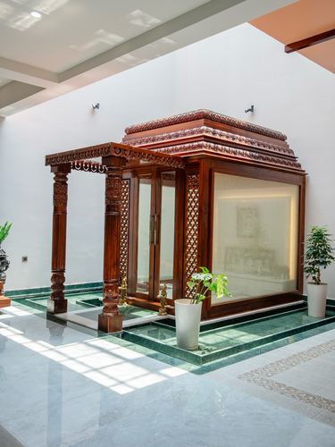 Carved wooden pooja pavilion with glass doors on green stone platform in sunlit atrium