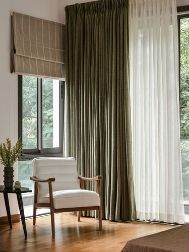 Cream boucle armchair beside floor-to-ceiling olive curtains and sheer drapes near a garden-view window