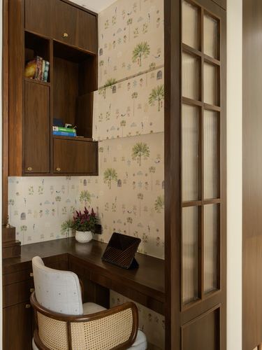 Wooden study nook with patterned wallpaper, open shelves, cane chair and reeded glass door