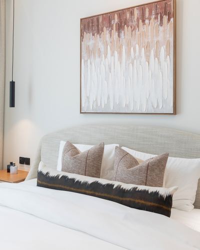 Neutral bedroom with upholstered headboard, textured artwork, and patterned lumbar pillow above white bedding