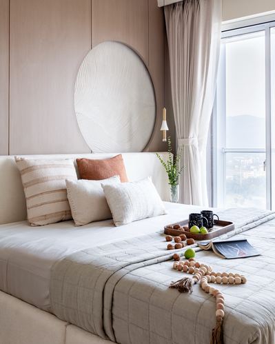Bed with layered neutral pillows below a round textured wall panel, city view through window
