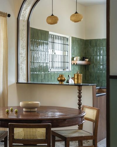 Dining area with round wood table and cane chairs opens through an arch to a green-tiled kitchen
