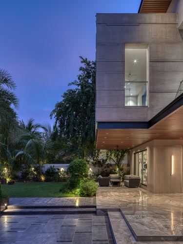Twilight facade of a modern villa with stone cladding, timber-lined overhang, and lit terrace beside a garden