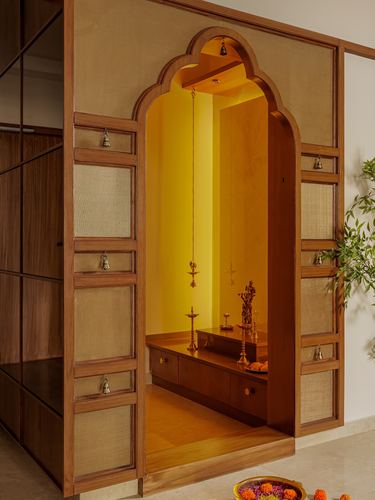 Wooden cusped-arch pooja room door with cane panels opening to a golden-lit shrine with brass idol and hanging bells