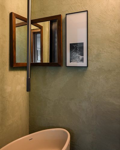 Textured olive plaster bathroom wall with wooden mirror, framed print, and freestanding tub corner