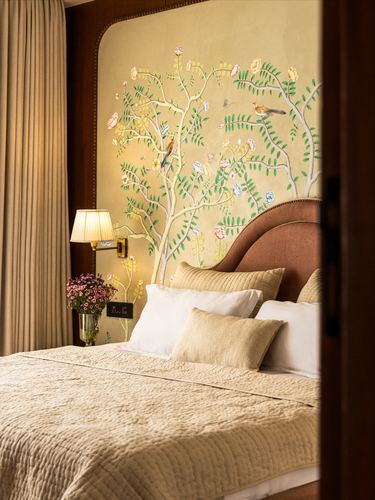 Bed with terracotta upholstered headboard against a hand-painted floral and bird mural, warm lamp light