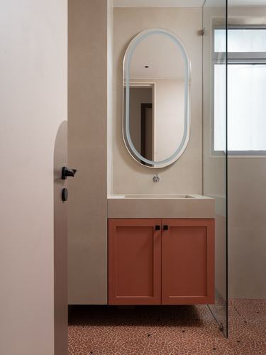 Bathroom vanity with oval backlit mirror and terracotta cabinet against beige stone wall