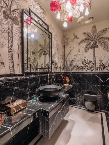 Powder room with black marble vanity, round basin, and tropical mural wallpaper under a colorful glass chandelier