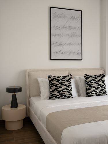 Neutral bedroom with upholstered headboard, patterned cushions, abstract art, and dark table lamp on round nightstand