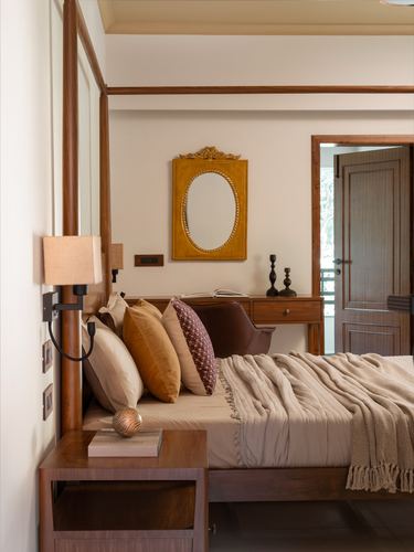 Four-poster wooden bed with layered cushions beneath an ornate oval mirror in a warm-toned bedroom