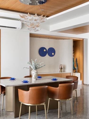Dining table with tan leather chairs under a sculptural chandelier, wood ceiling, blue wall art beyond