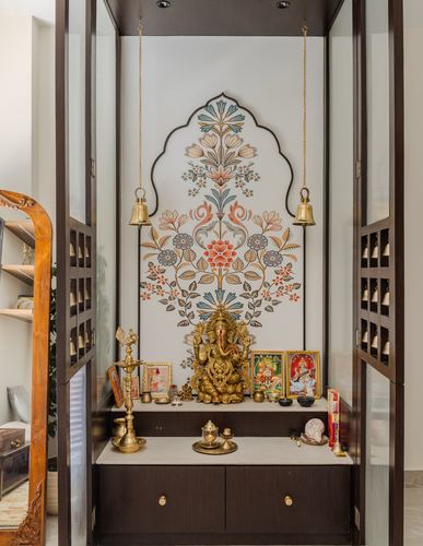 Wooden pooja unit with arched floral mural, brass idol, and hanging bells