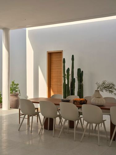 Outdoor dining table with white chairs beneath a covered patio beside cacti and a slatted wood door